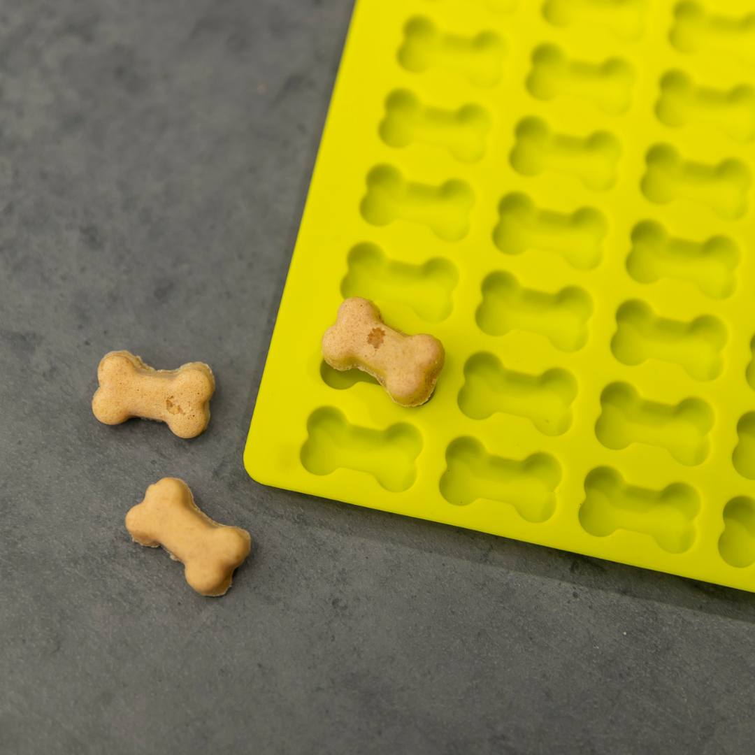 Selbstgebackene Hundeleckerlis in Knochenform neben gelber Silikon Backmatte für Hundekekse, frisch gebacken und natürlich.