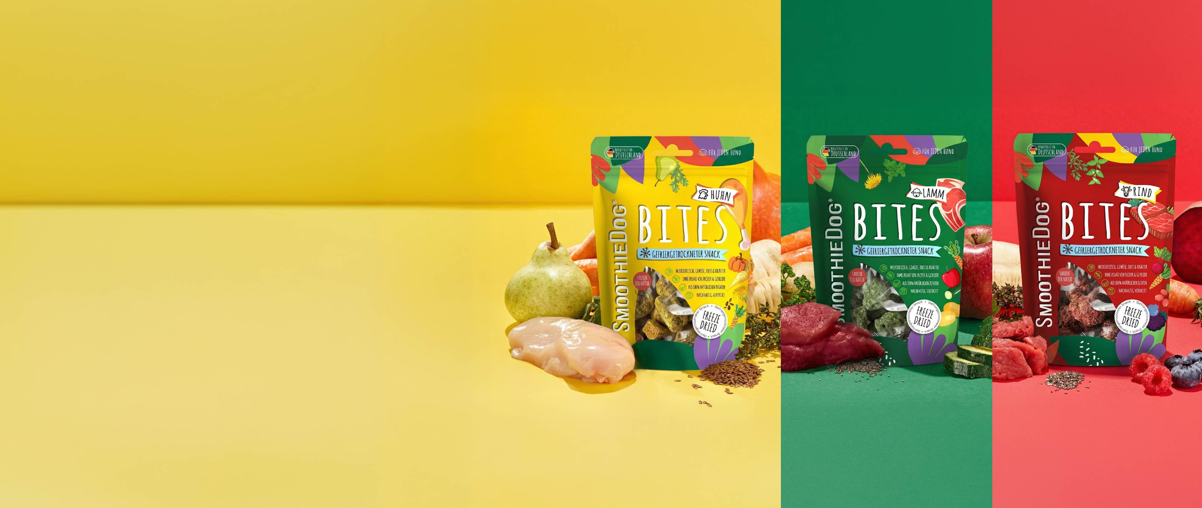Banner mit drei SmoothieDog Bites Sorten – Huhn, Lamm und Rind – jeweils in farblich abgestimmten Hintergründen (gelb, grün, rot) dargestellt. Vor den Verpackungen liegen die passenden Zutaten wie Fleisch, Gemüse, Obst und Kräuter, die die natürlichen Inhaltsstoffe der gefriergetrockneten Hundesnacks zeigen.