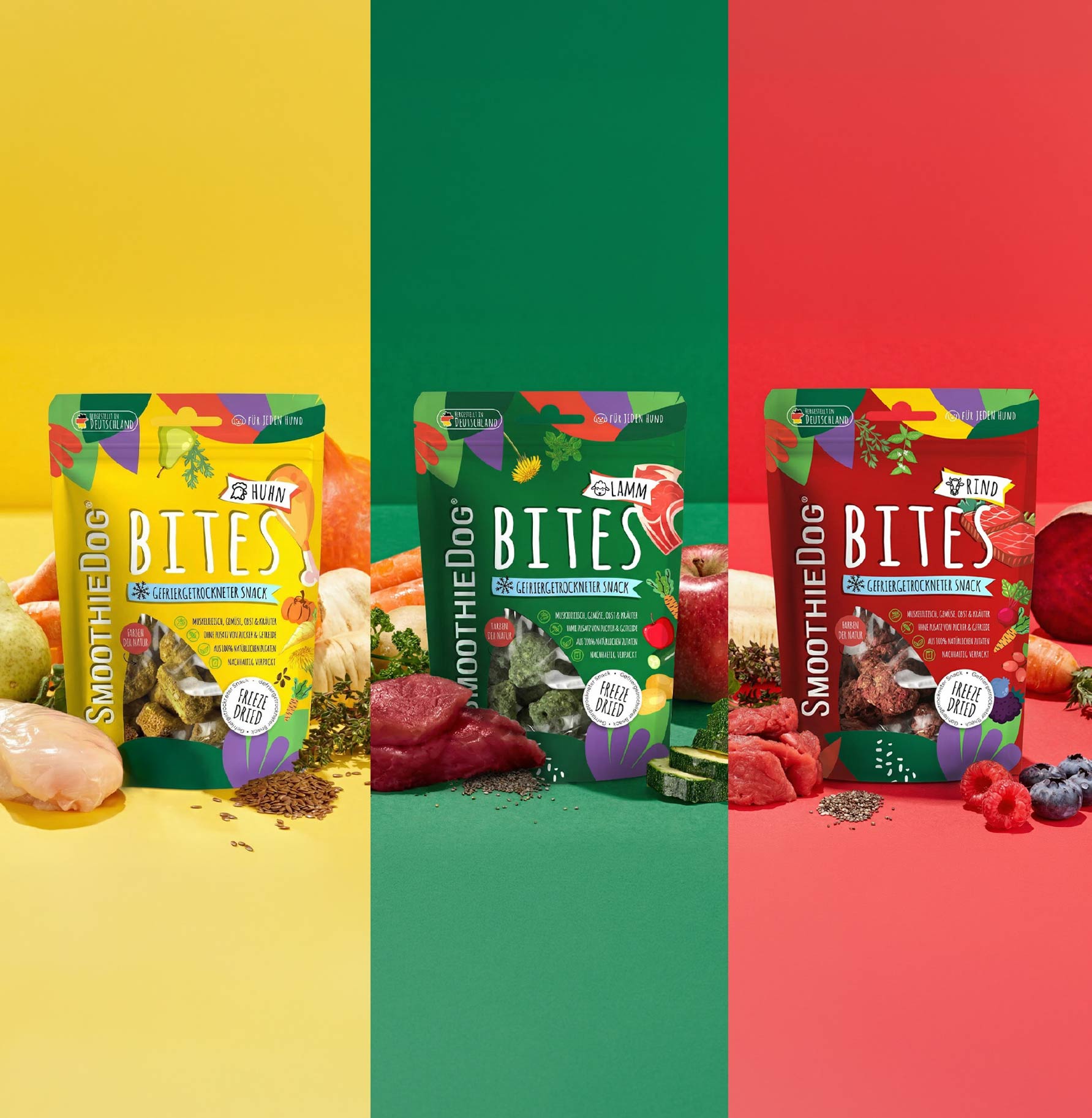 Banner mit drei SmoothieDog Bites Sorten – Huhn, Lamm und Rind – jeweils in farblich abgestimmten Hintergründen (gelb, grün, rot) dargestellt. Vor den Verpackungen liegen die passenden Zutaten wie Fleisch, Gemüse, Obst und Kräuter, die die natürlichen Inhaltsstoffe der gefriergetrockneten Hundesnacks zeigen.