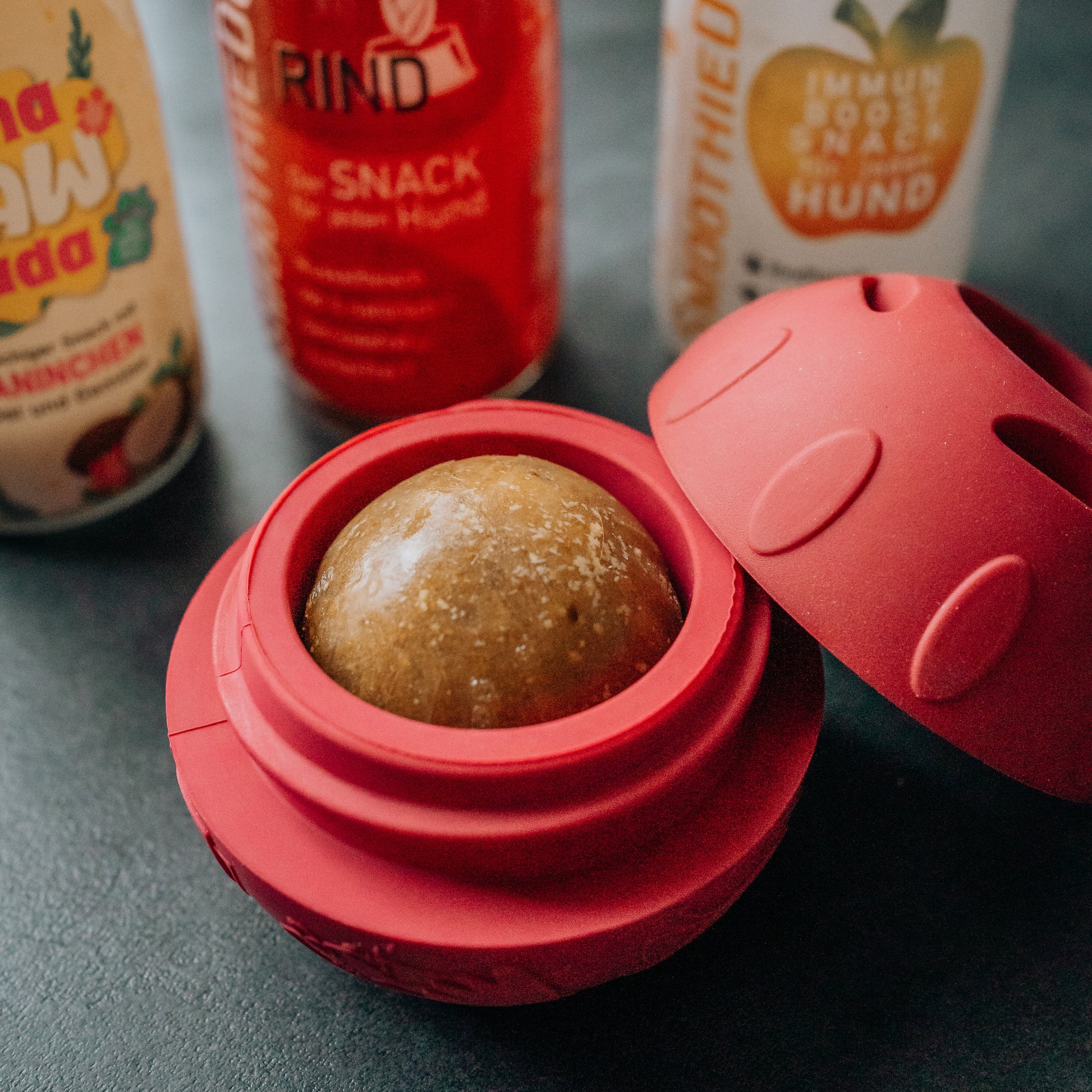 SmoothieDog Schleckball befüllt mit gefrorenem Snack: Der rote Schleckball ist geöffnet und enthält eine gefrorene Kugel als Snackfüllung. Im Hintergrund stehen verschiedene SmoothieDog Flaschen. Das Bild zeigt, wie der Schleckball für die Hundebeschäftigung genutzt wird.